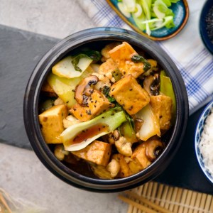 Soy tofu and vegetable stir-fry in a black pot topped with sesame seeds, on a dining table with side dishes and chopsticks.