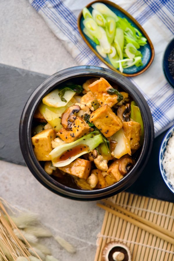 Soy tofu and vegetable stir-fry in a black pot topped with sesame seeds, on a dining table with side dishes and chopsticks.