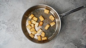 Skillet with several golden-brown, bite-sized tofu cubes sautéed in oil on a gray countertop