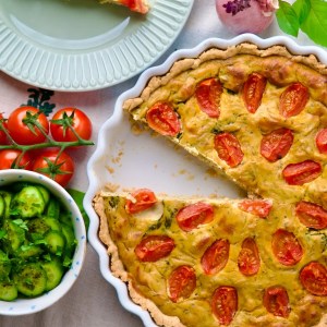 Quiche with cherry tomato halves and herbs in a white fluted tart dish, a slice on a pale green plate nearby.