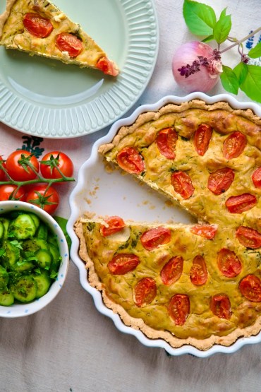 Quiche with cherry tomato halves and herbs in a white fluted tart dish, a slice on a pale green plate nearby.