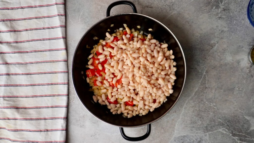 Top-down view of a black pot filled with white beans and diced red peppers cooking on a gray countertop, seen from above.